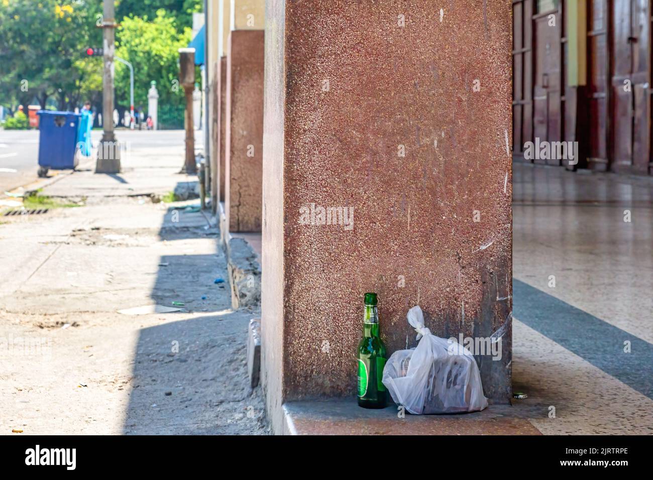 Eine Plastiktüte mit Müll und eine Bierflasche werden auf der Veranda des Gebäudes abgelegt. In der Ecke sind große Mülltonnen der Stadt zu sehen. Stockfoto