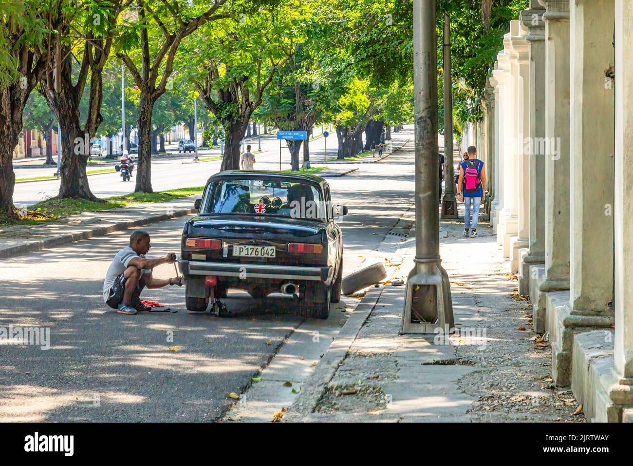 Ein Kubaner wechselt den Reifen eines russischen Fahrzeugs auf einer Stadtstraße. Der Bürgersteig rechts ist beschädigt oder nicht gewartet. Stockfoto