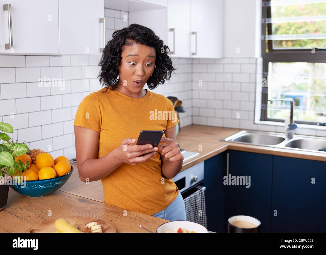 Eine junge Frau sieht schockiert aus, als sie beim Frühstück in der Küche auf ihrem Telefon scrollt Stockfoto