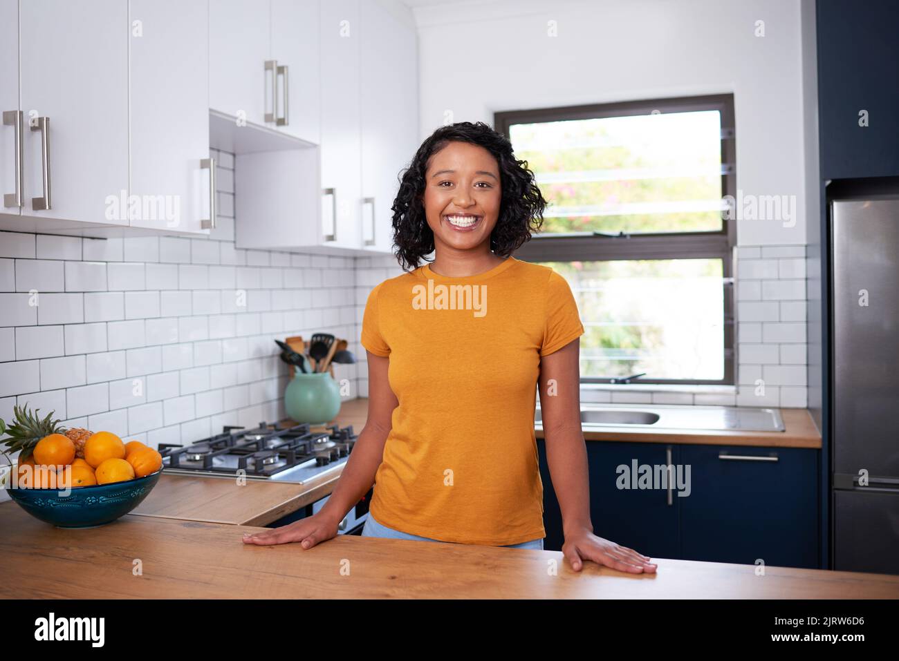 Eine junge, multiethnische Frau lächelt und steht in einer kleinen, sauberen Küche Stockfoto