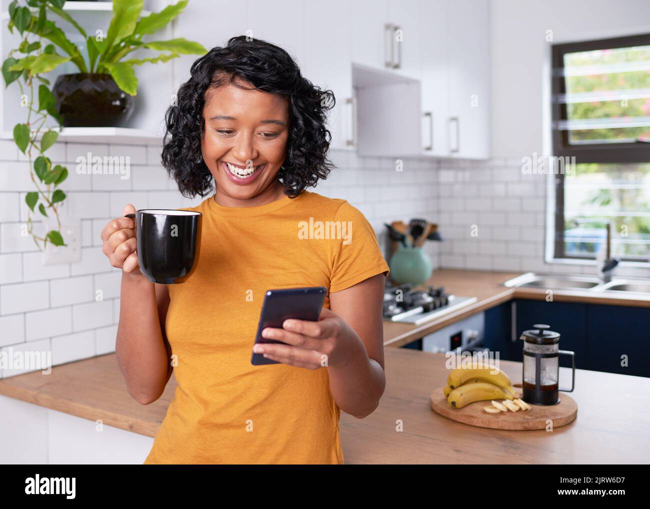 Eine junge, multiethnische Frau lacht und schaut in der Küche auf ihr Telefon Stockfoto