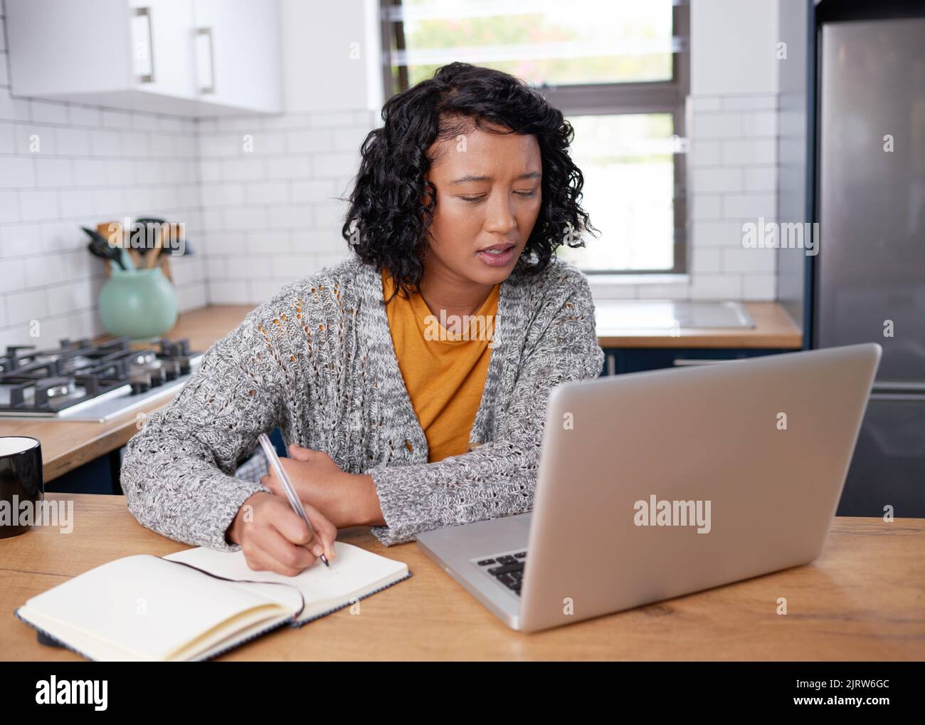 Eine junge, multiethnische Frau arbeitet von zu Hause aus in der Küche und notiert sich Stockfoto