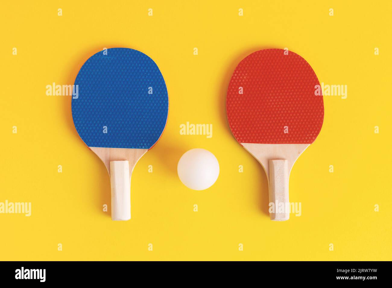 Ein Paar blaue und rote Tischtennisschläger und weiße Kugel auf gelbem Hintergrund. Draufsicht, Studioaufnahme. Tischtennis- oder Tischtennisausrüstung. Stockfoto