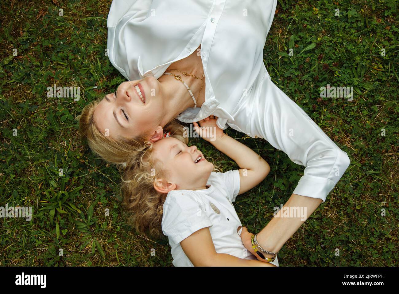 Draufsicht auf lachende Familie mit geschlossenen Augen, die im Sommer auf Gras liegen und Spaß haben. Junge Frau, die sich mit einem kleinen Mädchen entspannt. Stockfoto