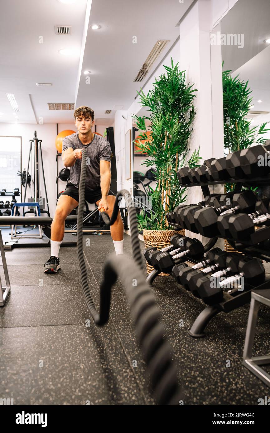 Ein junger männlicher Athlet, der tagsüber im modernen Fitnessstudio trainieren kann, mit Kampfseilen in der Nähe des Hantelracks Stockfoto