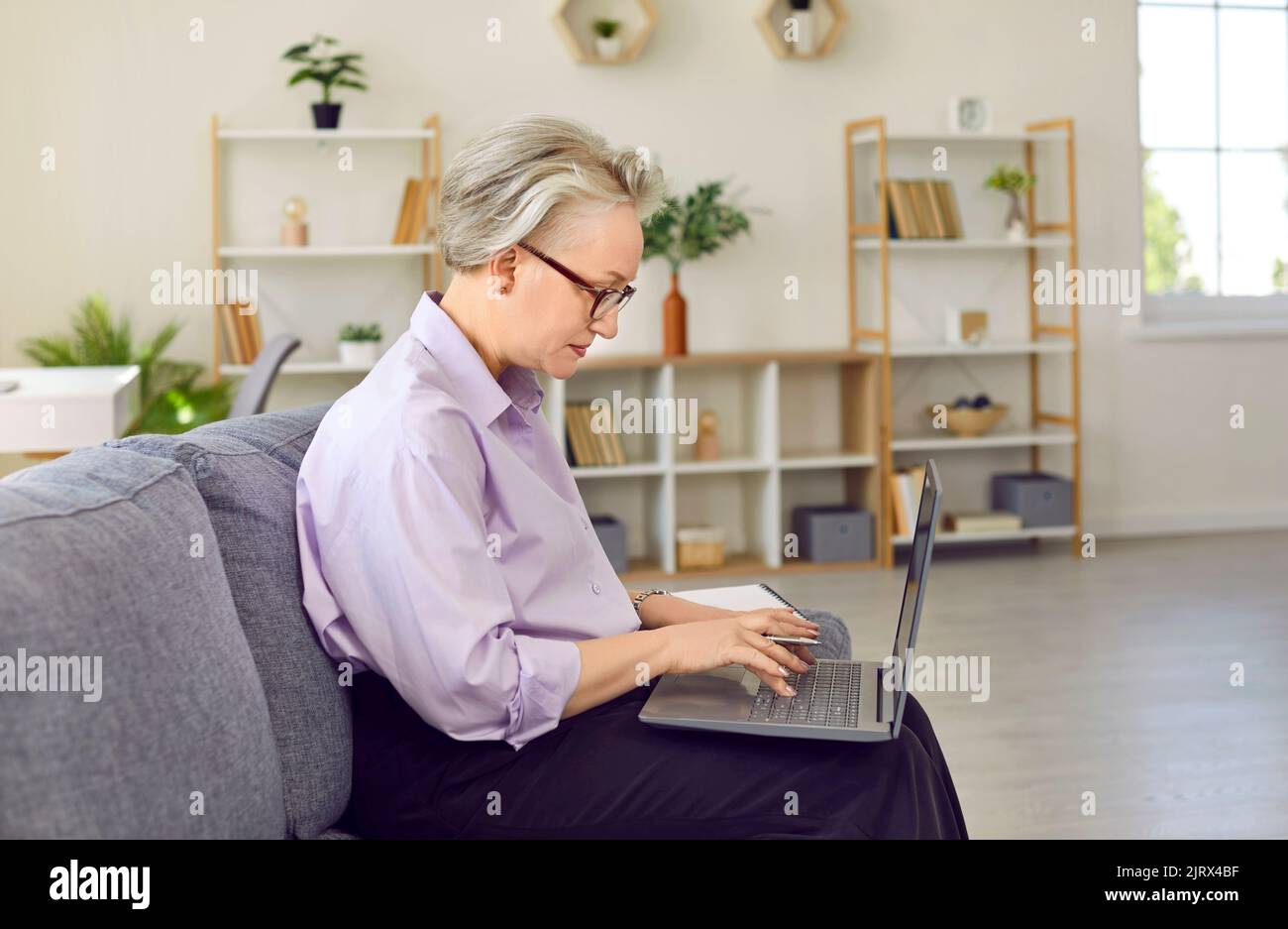 Ältere erfolgreiche Frau mit grauen Haaren arbeitet sitzt auf der Couch mit Laptop auf den Knien Stockfoto