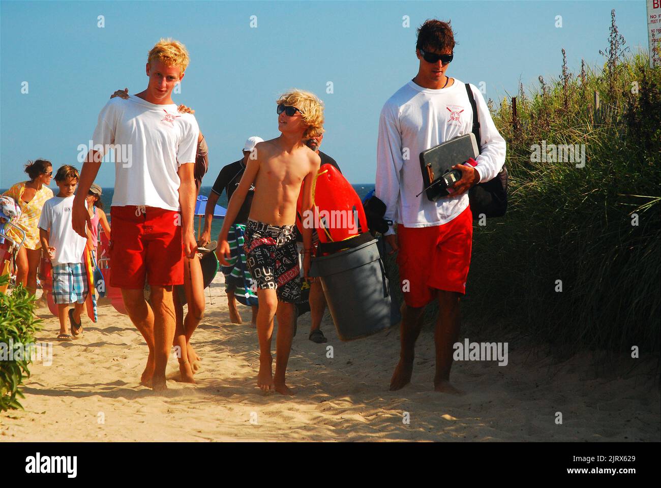 Ein kleiner Junge geht spazieren und spricht mit zwei Rettungsschwimmern, während sie den Strand verlassen und ihre Ausrüstung an einem Sommerferientag am Strand tragen Stockfoto