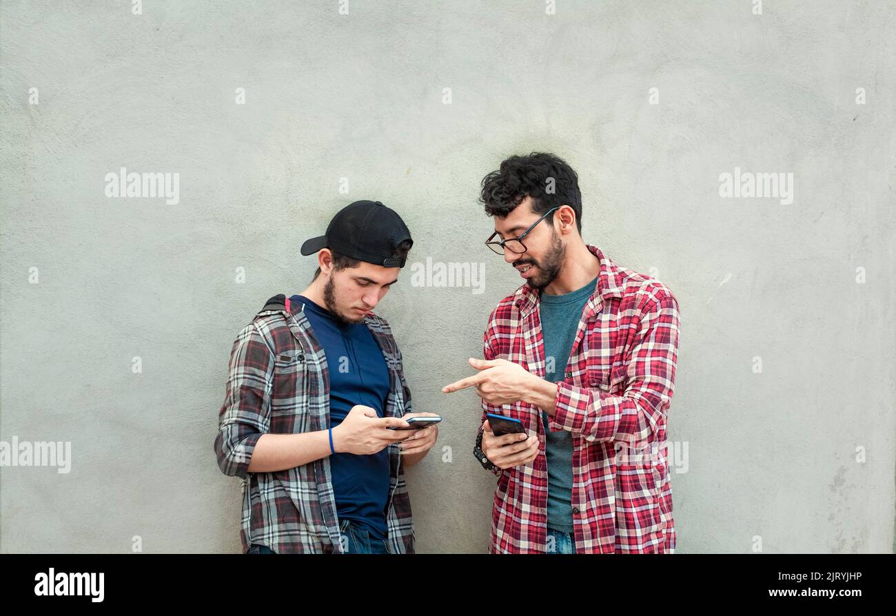 Zwei junge Freunde, die sich an einer Wand lehnten, um ihre Handys zu überprüfen, zwei Freunde, die sich an einer Wand lehnten, um die Inhalte auf ihren Handys zu sehen. Stockfoto