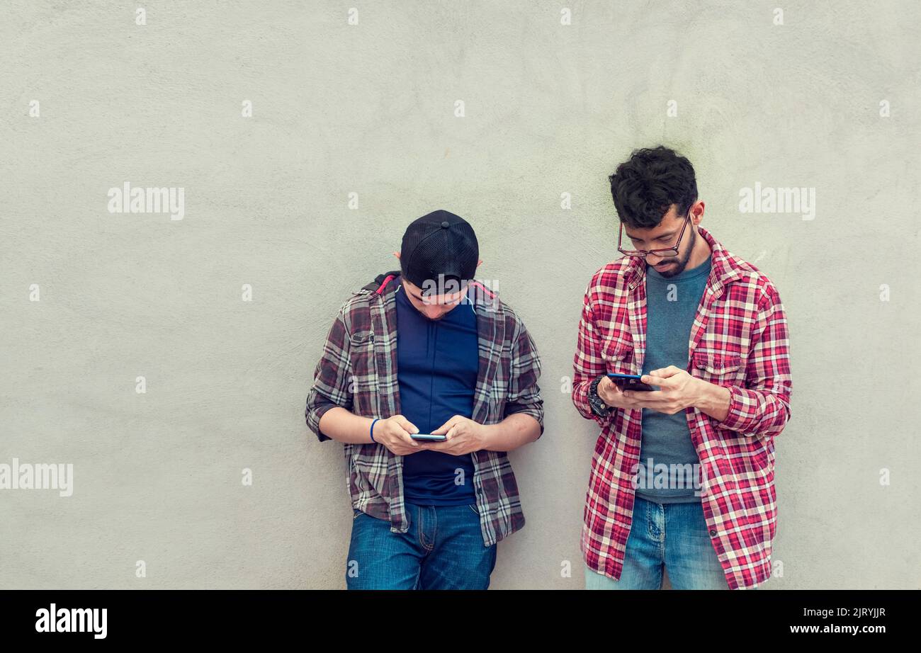 Zwei Teenager-Freunde an einer Wand, die ihre Handys überprüfen, zwei Freunde, die sich an einer Wand lehnen und auf ihren Telefonen SMS schreiben. Freund, der ihm das Handy zeigt Stockfoto