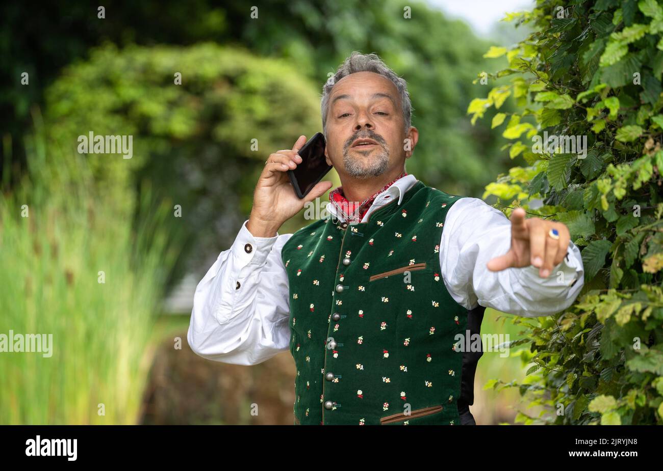 Mann mit Telefon in bayerischer Tracht an einer Hecke, Karlsruhe, Deutschland Stockfoto