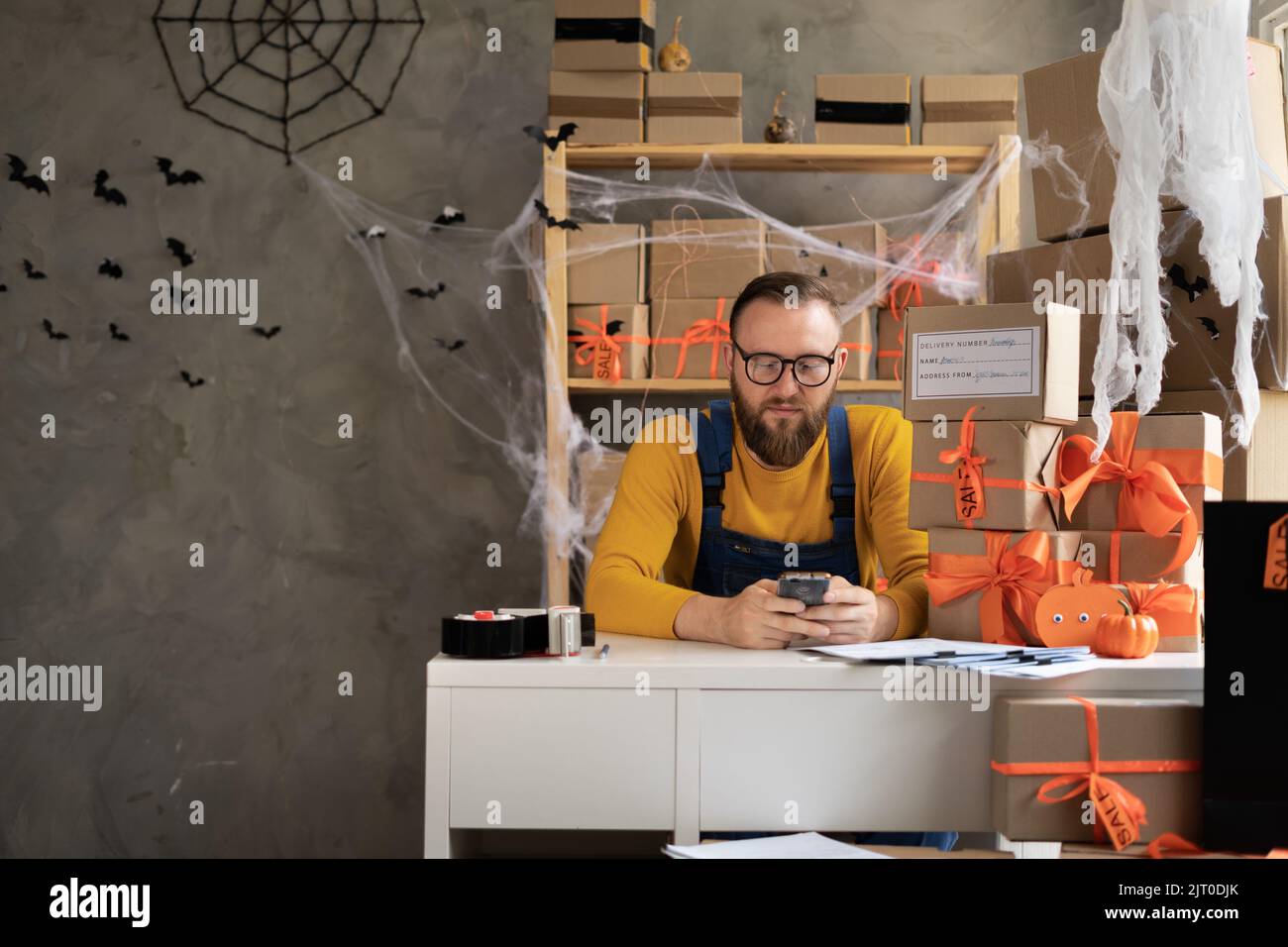 Der Besitzer eines Kleinunternehmens verwendet eine mobile App auf dem Smartphone, während er das Paket überprüft. Lagerarbeiter, Verkäufer in Brillen, der ein Telefon hält. Großer Verkauf Stockfoto