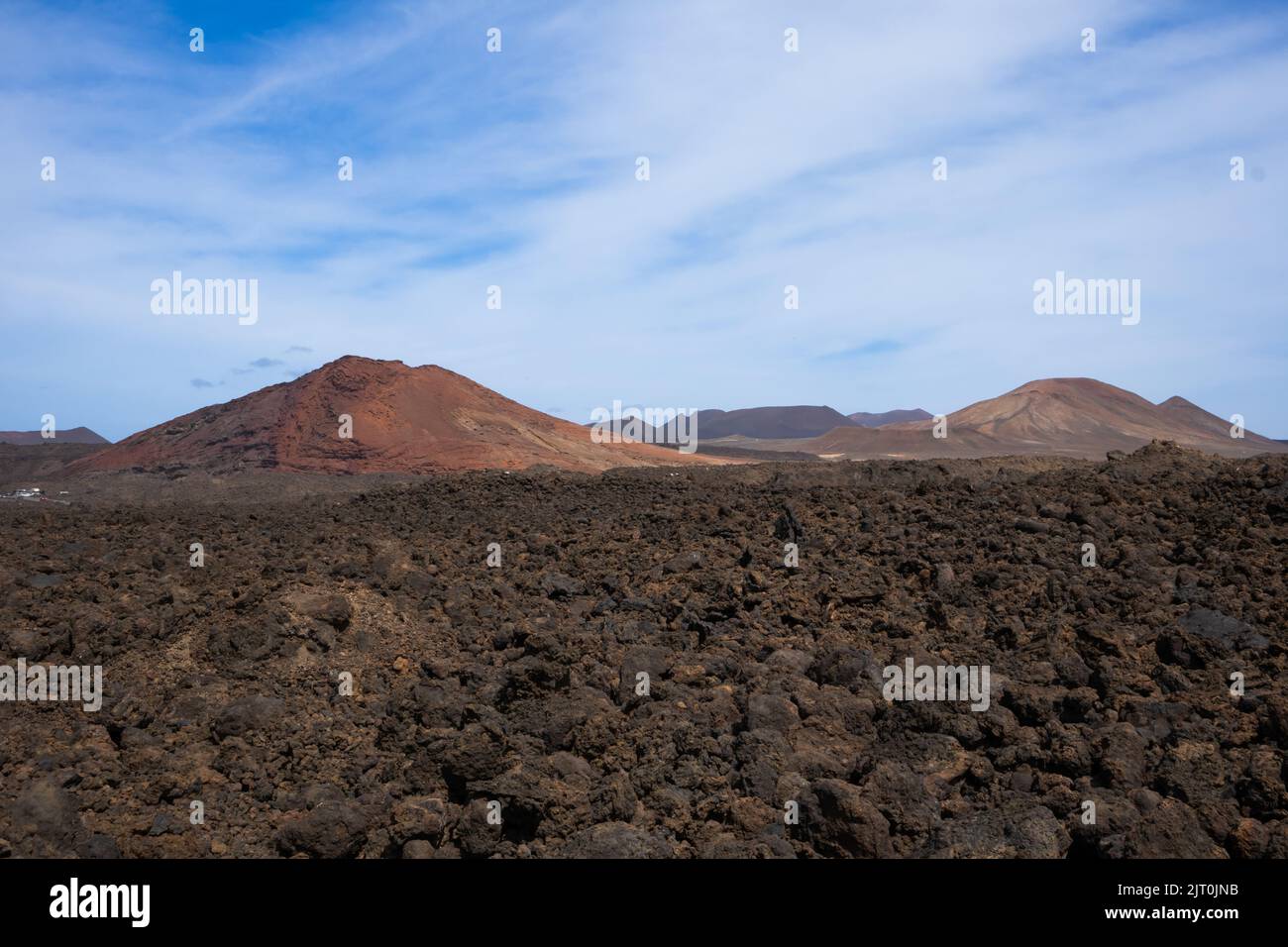 Lavaboden von brauner bis schwarzer Farbe. Berge im Hintergrund. Blauer Himmel mit weißen Wolken im frühen Frühjahr. Lanzarote, Las Palmas, Kanarische Inseln Stockfoto