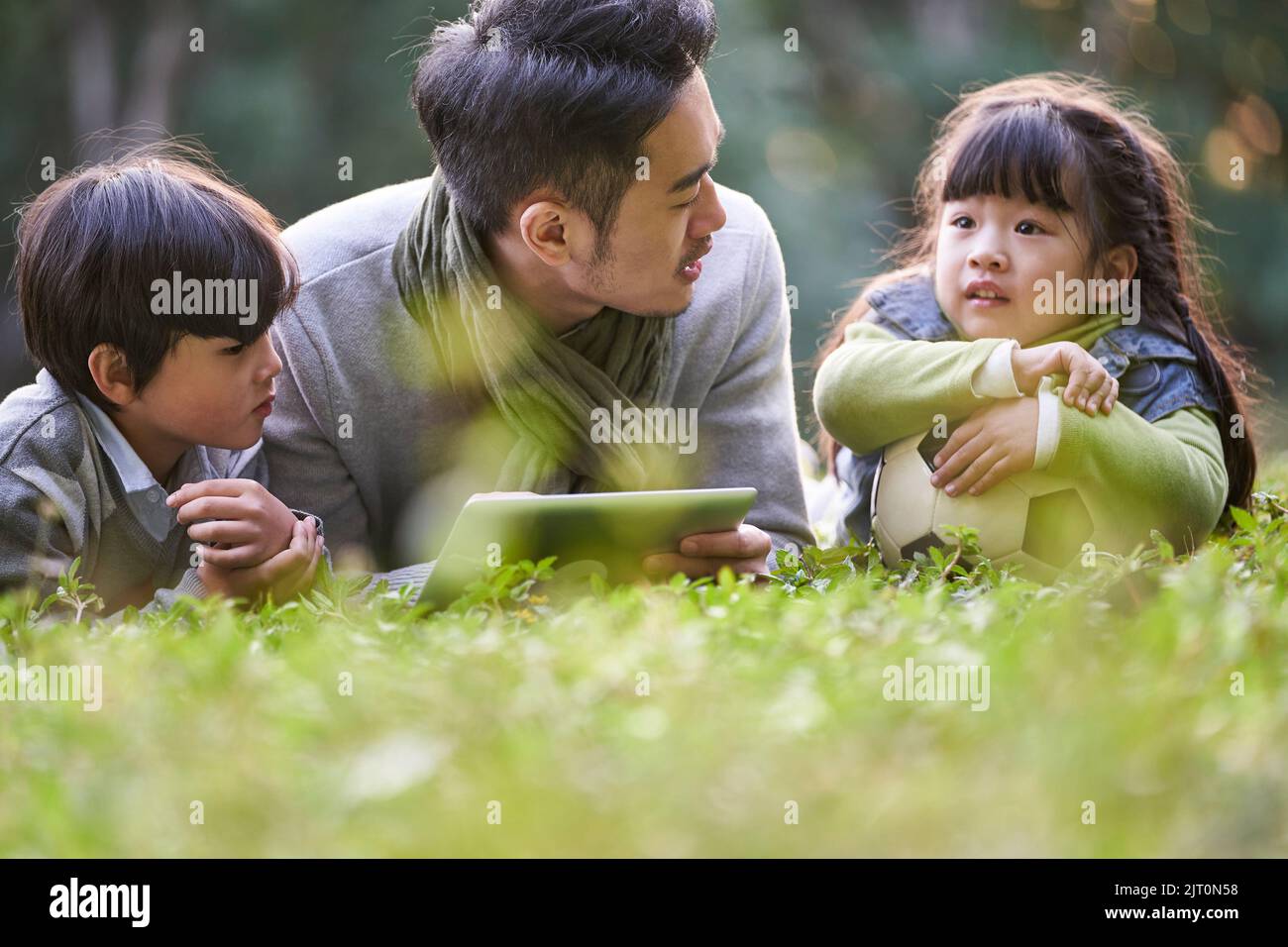 asiatischer Vater, der vorne auf Gras liegt und zwei Kindern glücklich und lächelnd Geschichten erzählt Stockfoto