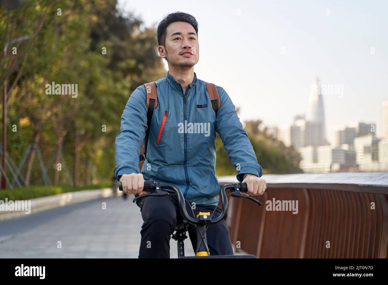 Junger asiatischer Erwachsener, der im Park am Flussufer mit der Skyline der Stadt im Hintergrund ein Fahrrad fährt Stockfoto