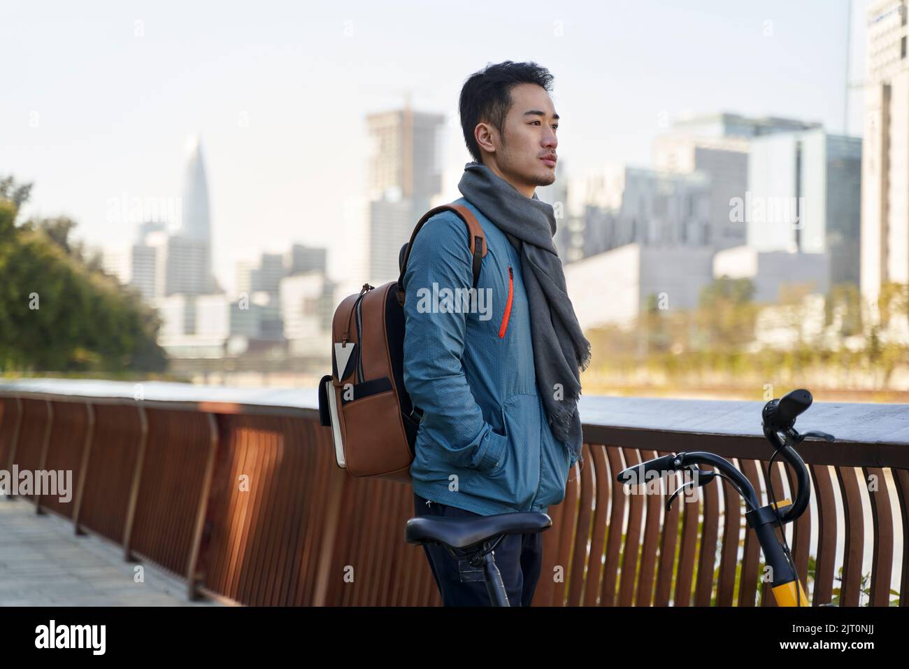Junger asiatischer Erwachsener mit Rucksack, der an seinem Fahrrad steht und die Skyline der Stadt im Park am Flussufer betrachtet Stockfoto