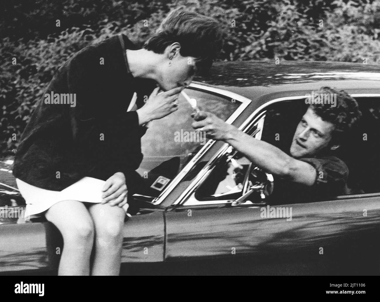 In der 1960s. Eine Frau sitzt auf der Motorhaube eines Autos, und ein Mann greift mit einem Zigarettenanzünder, um sie anzuzünden. Das Jahr ist 1964. Schweden Stockfoto
