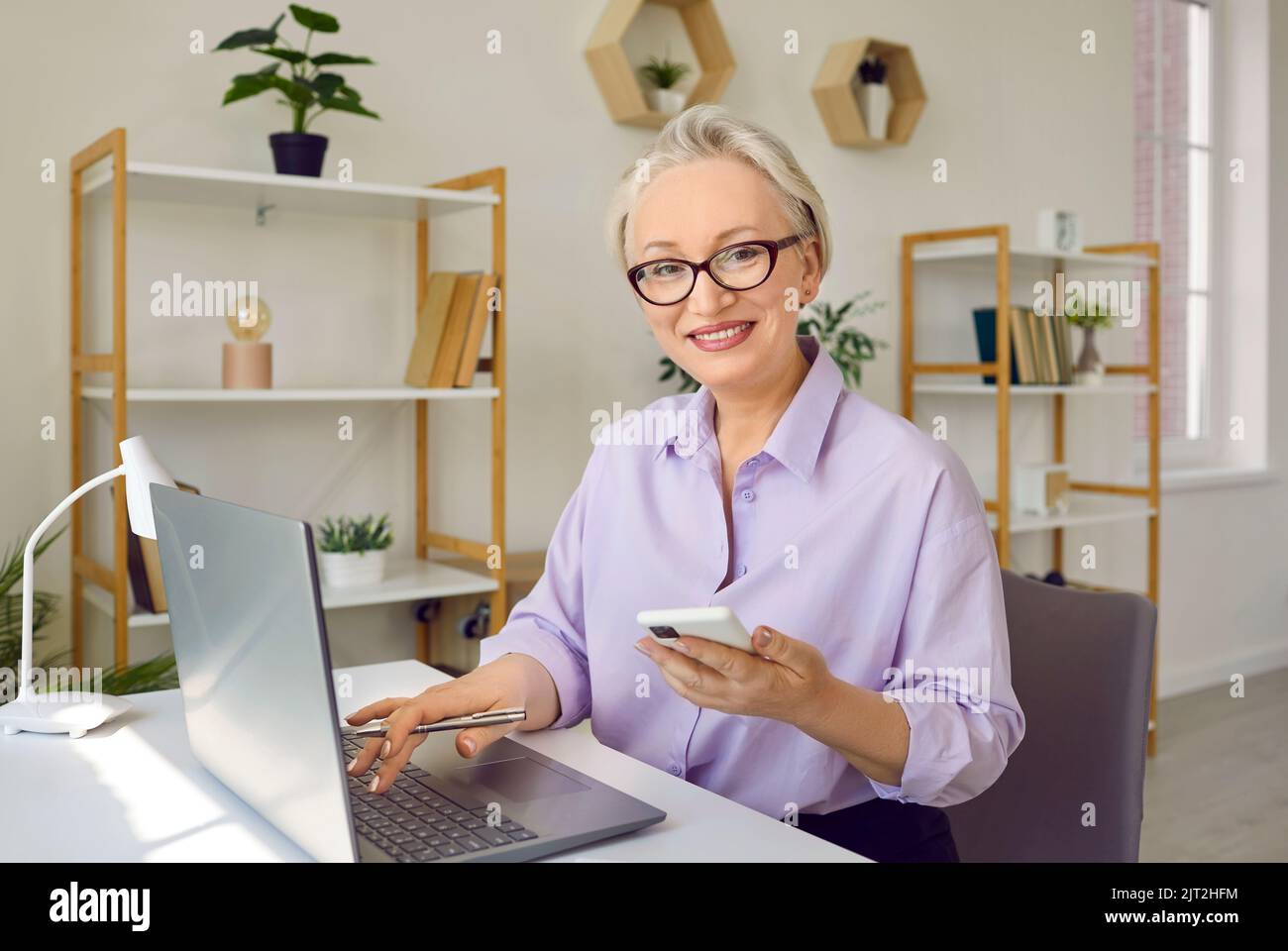 Eine schöne lächelnde ältere Geschäftsfrau, die bei ihrer Arbeit Handy und Laptop benutzte. Stockfoto
