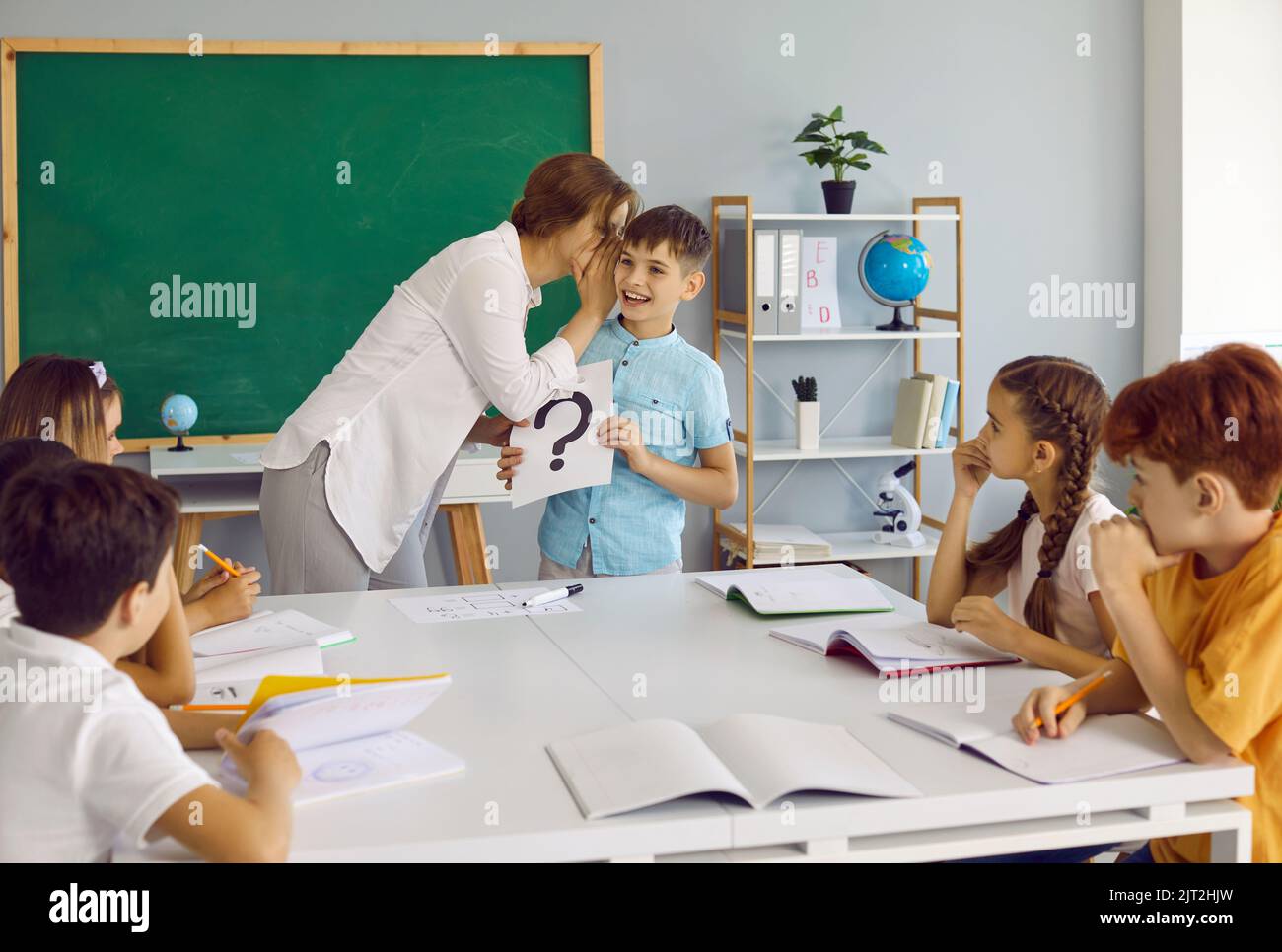 Die freundliche Lehrerin flüstert dem kleinen Schuljungen zu Ohren, der während des Schulunterrichts Hinweise gibt. Stockfoto