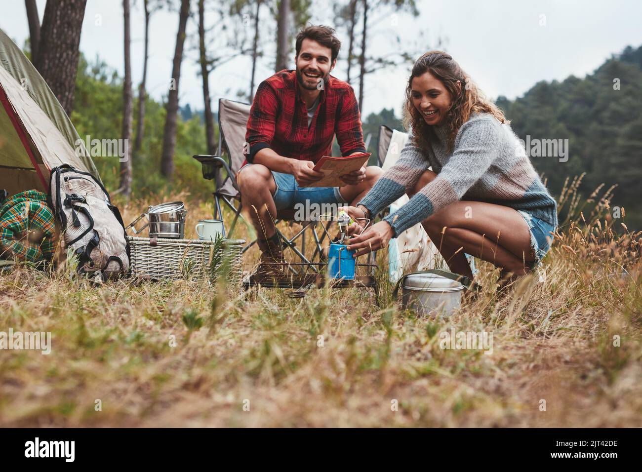 Glückliches junges Paar Camping im Freien in der Natur. Eine fröhliche junge Frau zündet einen Lagerofen an, während ihr Freund dabeisitzt. Stockfoto