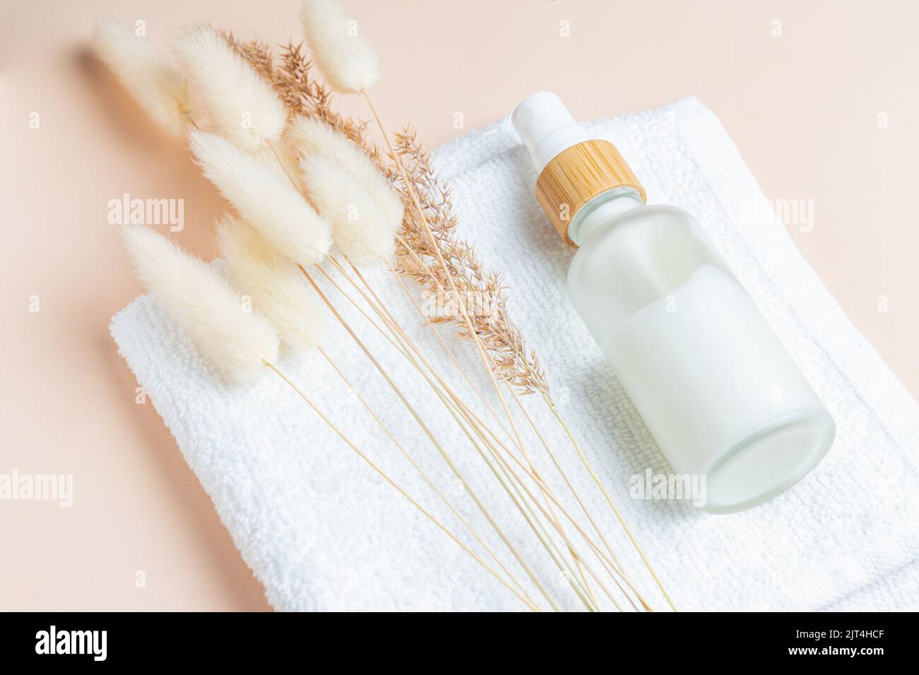 Milchglasflasche mit Gesichtscreme oder Flüssigkeit auf weißem Handtuch mit Lagurusblüten und trockenem Schilf. Hautpflege Öko-Konzept. Naturkosmetik Stockfoto