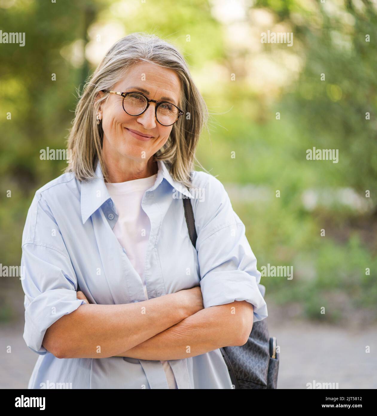 Schöne graue Haare reife Frau trägt Brille und Rucksack stehende Arme gefaltet im Freien genießen Urlaub rund um die Welt im Ruhestand reisen. Reife Frau mit perfekter Haut trägt blaues Hemd. Stockfoto