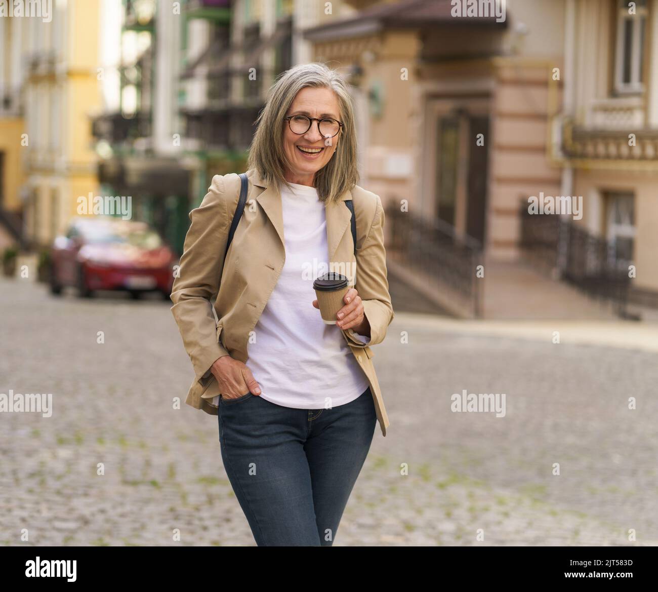 Aufgeregte reife Geschäftsfrau in legerer Jacke und Jeans halten Papiertasse mit heißem Kaffee draußen in der Altstadt. Ältere Frauen reisen in der europäischen Stadt und genießen eine Tasse Tee oder Kaffee. Stockfoto