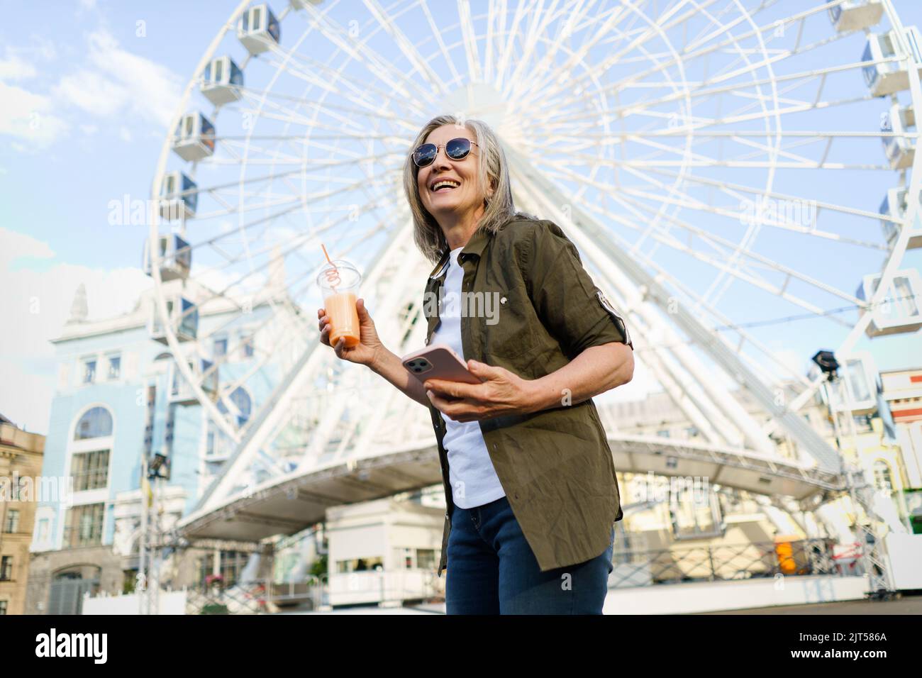 Fröhliche, grauhaarige europäische Frau, die gerne reist, hält Telefon, Smartphone, während sie unterwegs mit einer Plastikschale, Sonnenbrille und grünem Hemd in der Stadt Saft trinkt und auf dem Hintergrund ein Riesenrad hat. Stockfoto