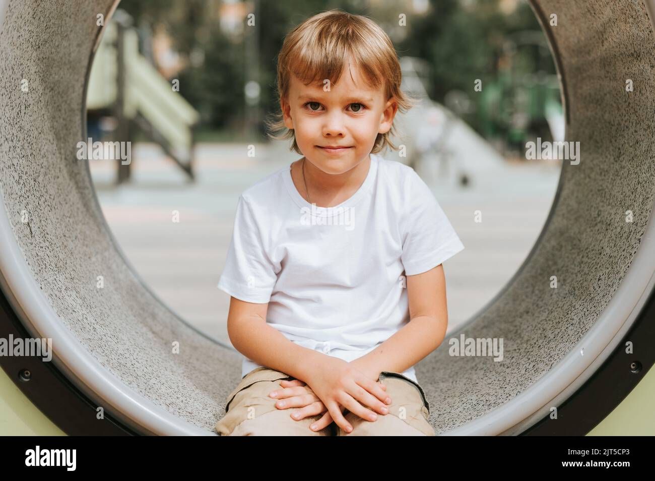 Kind spielt auf dem Stadtspielplatz auf Ring um. Nette kleine glücklich lächelnd offen fünf Jahre alten Jungen mit langen blonden Haaren in einem weißen T-Shirt. G Stockfoto