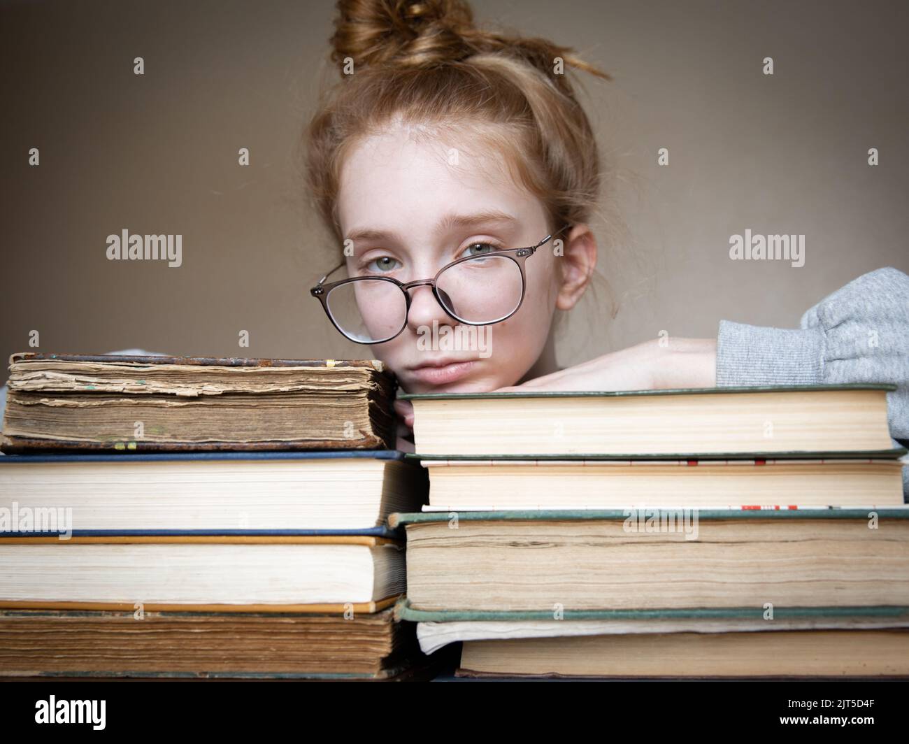 Auf dem Tisch liegen zwei große Stapel Bücher, auf denen ein Mädchen mit roten Haaren in einer Brille liegt. Das Mädchen hat einen traurigen Blick. Das Hauptobjekt des Buches. Stockfoto