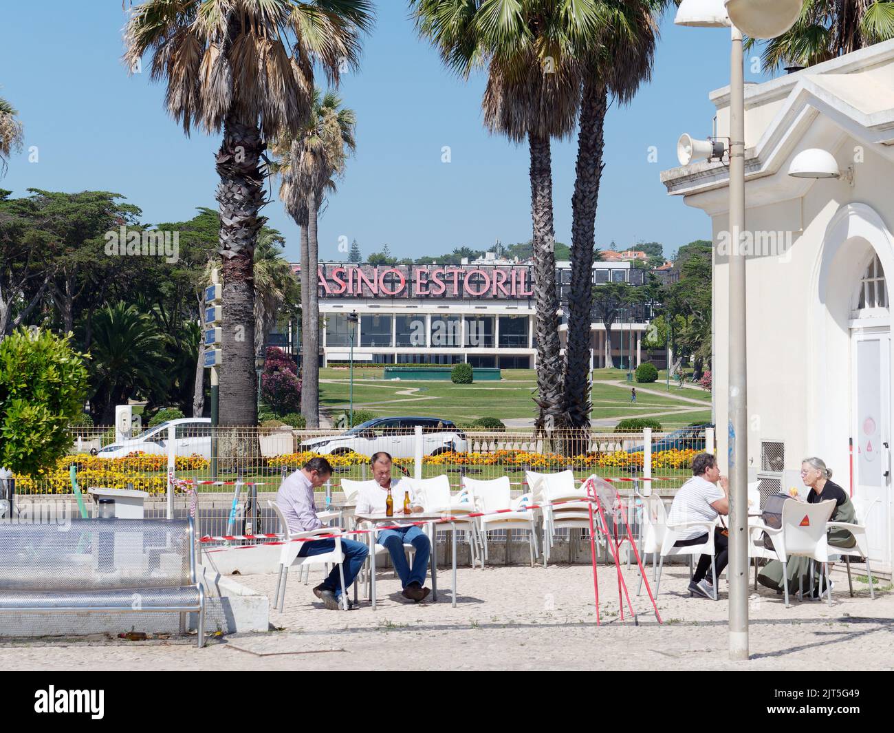 Estoril Casino hinter Palmen vom Bahnhof aus gesehen. Gemeinde Cascais, Portugal. Die Leute sitzen und trinken im Bahnhofscafé Stockfoto