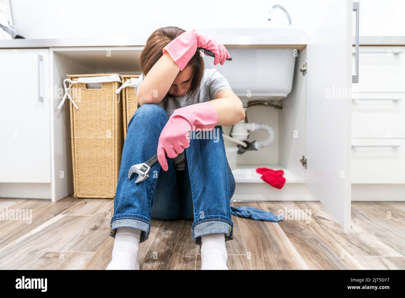 Traurige Frau, die in der Nähe des undichten Waschbeckens in der Waschküche mit einem verstellbaren Schraubenschlüssel saß Stockfoto