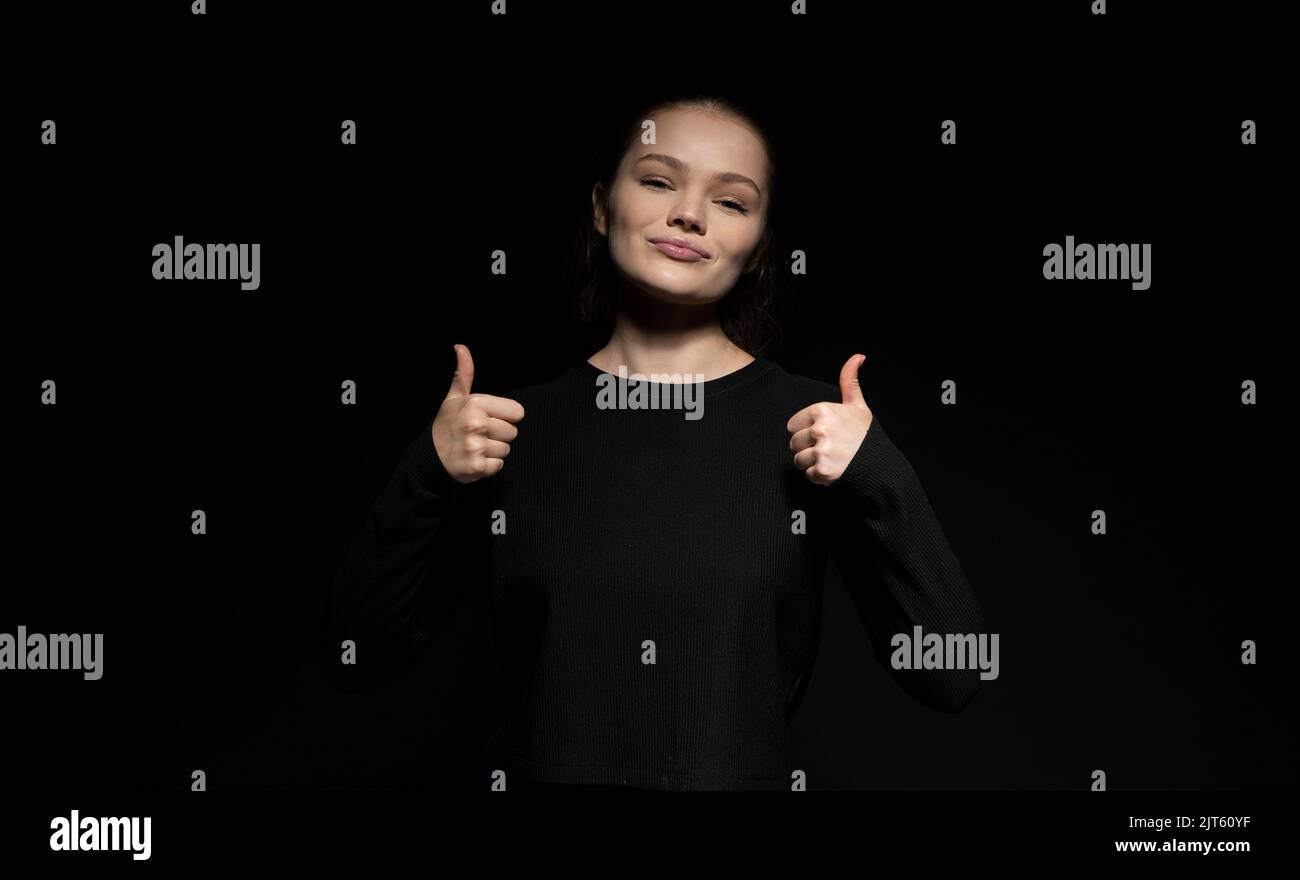 Junge fröhlich Brünette Frau trägt einen schwarzen Pullover mit Daumen über der schwarzen Wand. Stockfoto