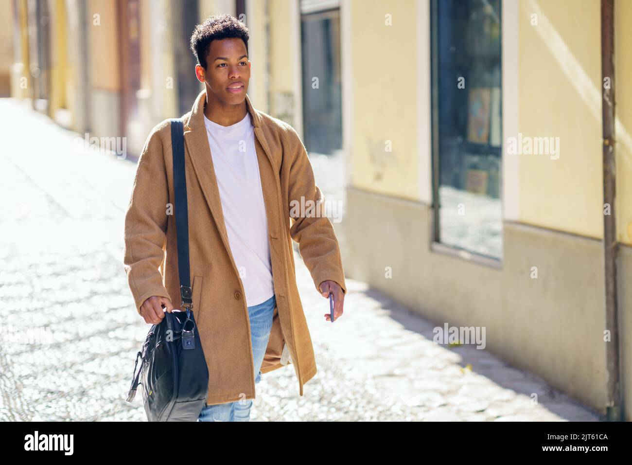 Ein junger schwarzer Mann, der die Straße entlang läuft, trägt eine Aktentasche und ein Smartphone. Stockfoto