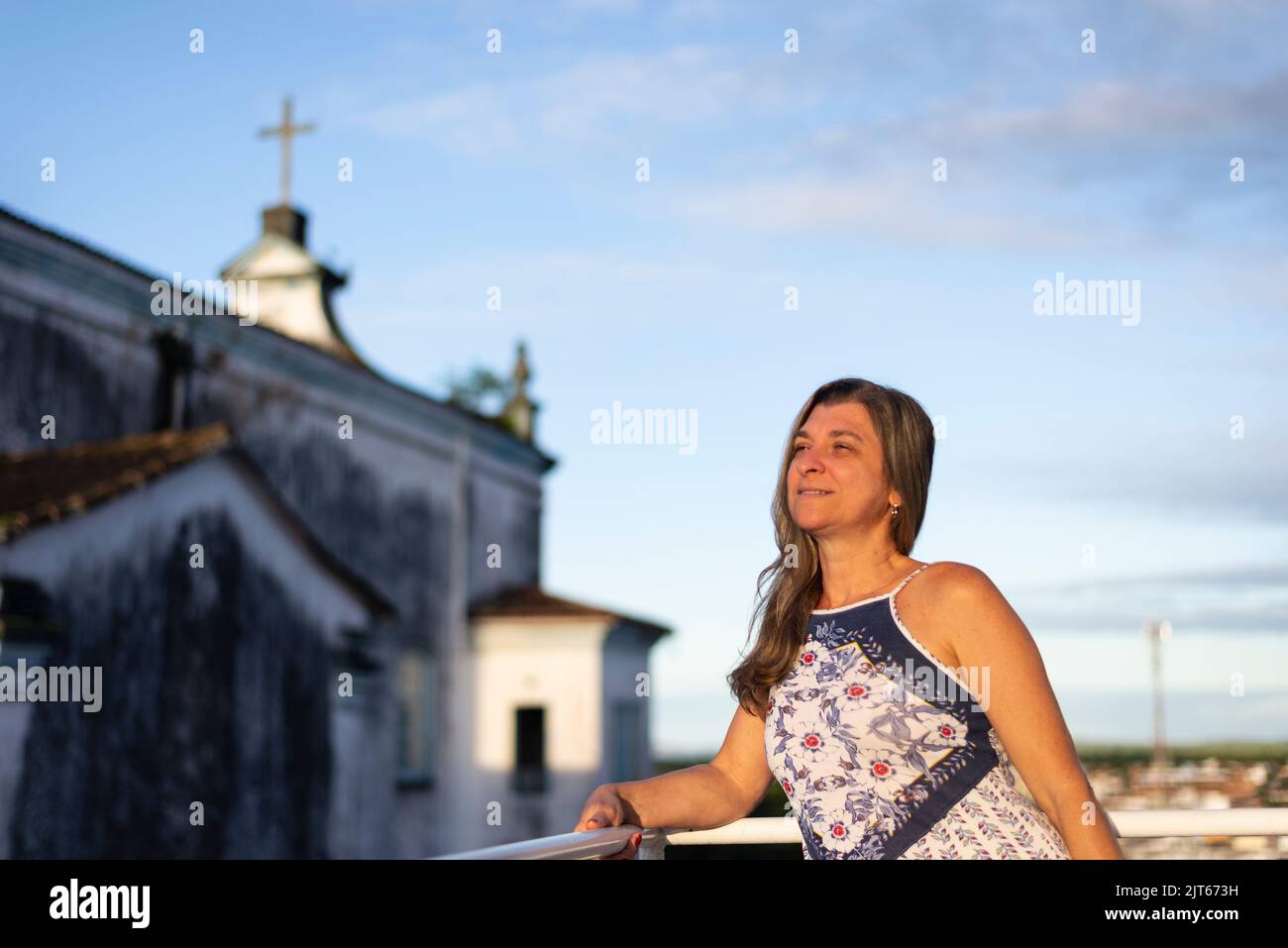 Eine Frau auf der Veranda ihres Hauses in hellen Kleidern blickt auf die Straße gegen den Himmel und die Kirche im Hintergrund. Stadt Valença, Bahia, Brasilien. Stockfoto