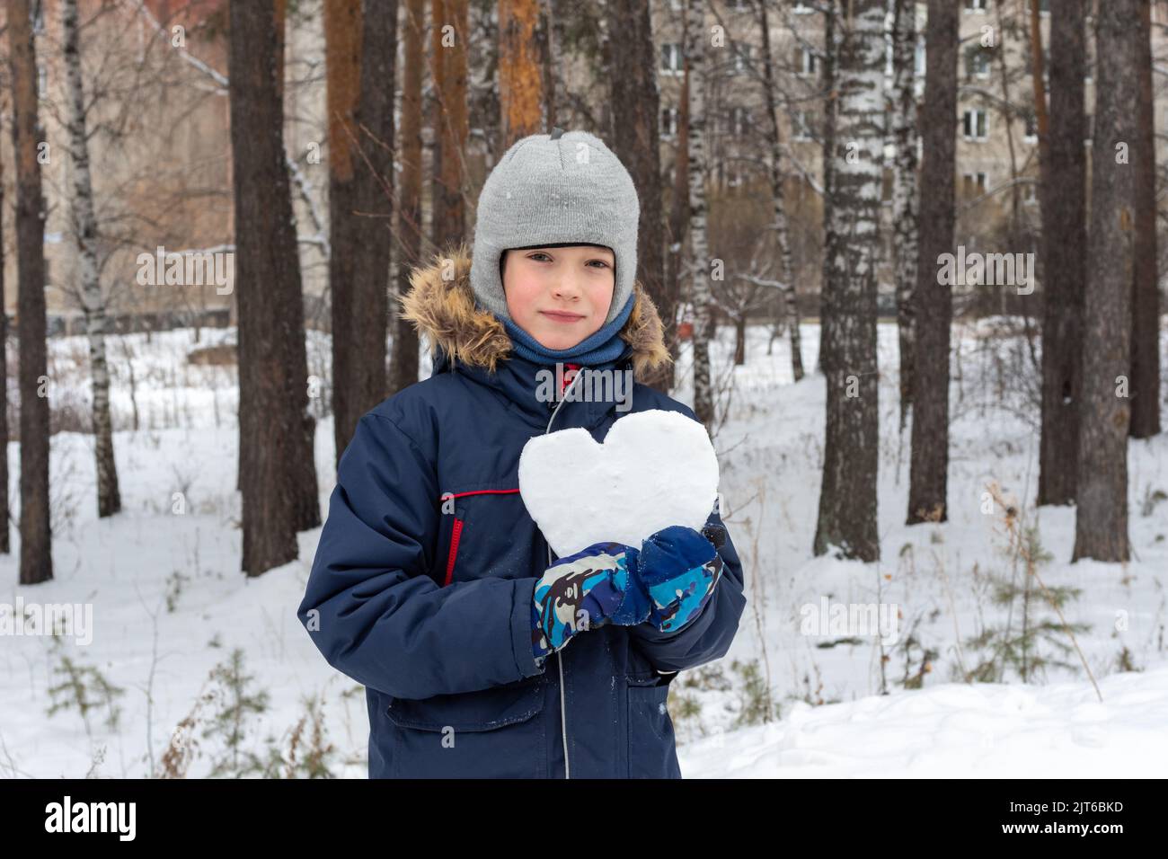 Kinder Winter. Niedlicher lächelnder Junge in einer Winterjacke und Fäustlingen, die nach einem Schneefall eine verschneite Herzform auf der Straße halten. Schnee Herz in den Händen. Stockfoto