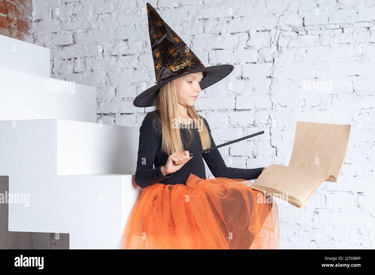 Halloween Für Kinder. Ein schönes, süßes Mädchen in einem Hexenkostüm, das einen Hut trägt, ein Buch mit Zaubersprüchen und einem Zauberstab hält, zaubert auf der Treppe sitzend. Stockfoto