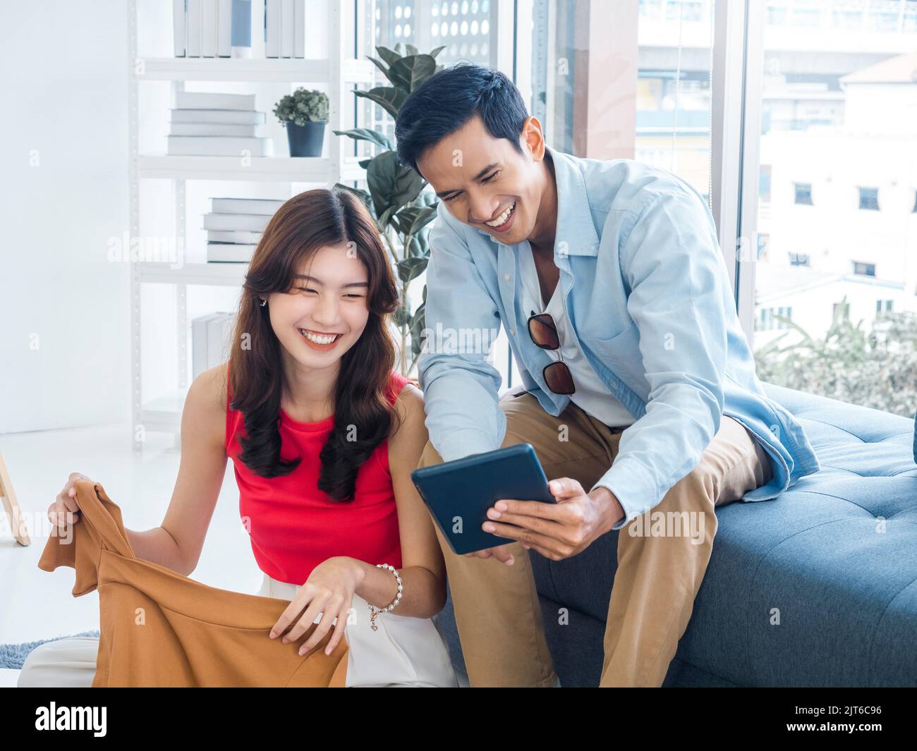 Glücklich schöner asiatischer Mann und junge schöne Frau lachen zusammen, während sie auf den Tablet-Bildschirm schauen. Frau hält ein Hemd und Mann hält Tablet auf Couc Stockfoto