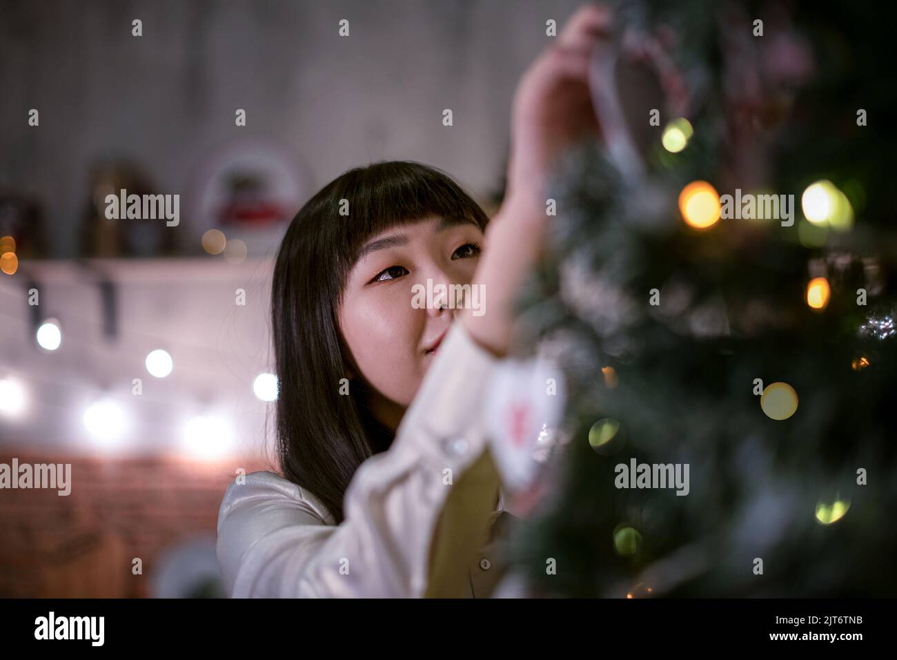 Ein hübsches dunkelhaariges koreanisches Mädchen in einem gemütlichen Weihnachtszimmer mit festlichen Bokeh-Lichtern am Abend kleidet einen Weihnachtsbaum mit Öko-natürlichem Spielzeug. Stockfoto