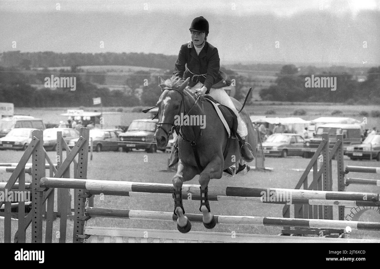 1980s, eine junge Frau, die bei einer County Show auf einem Pferd einen Zaun springt, Yorkshire, England, Großbritannien. Das Springreiten ist ein Reiterevent, bei dem Reiter und Pferd versuchen, über alle Zäune eines Parcours zu springen, ohne sie innerhalb einer festgelegten Frist niederzuschlagen. Stockfoto