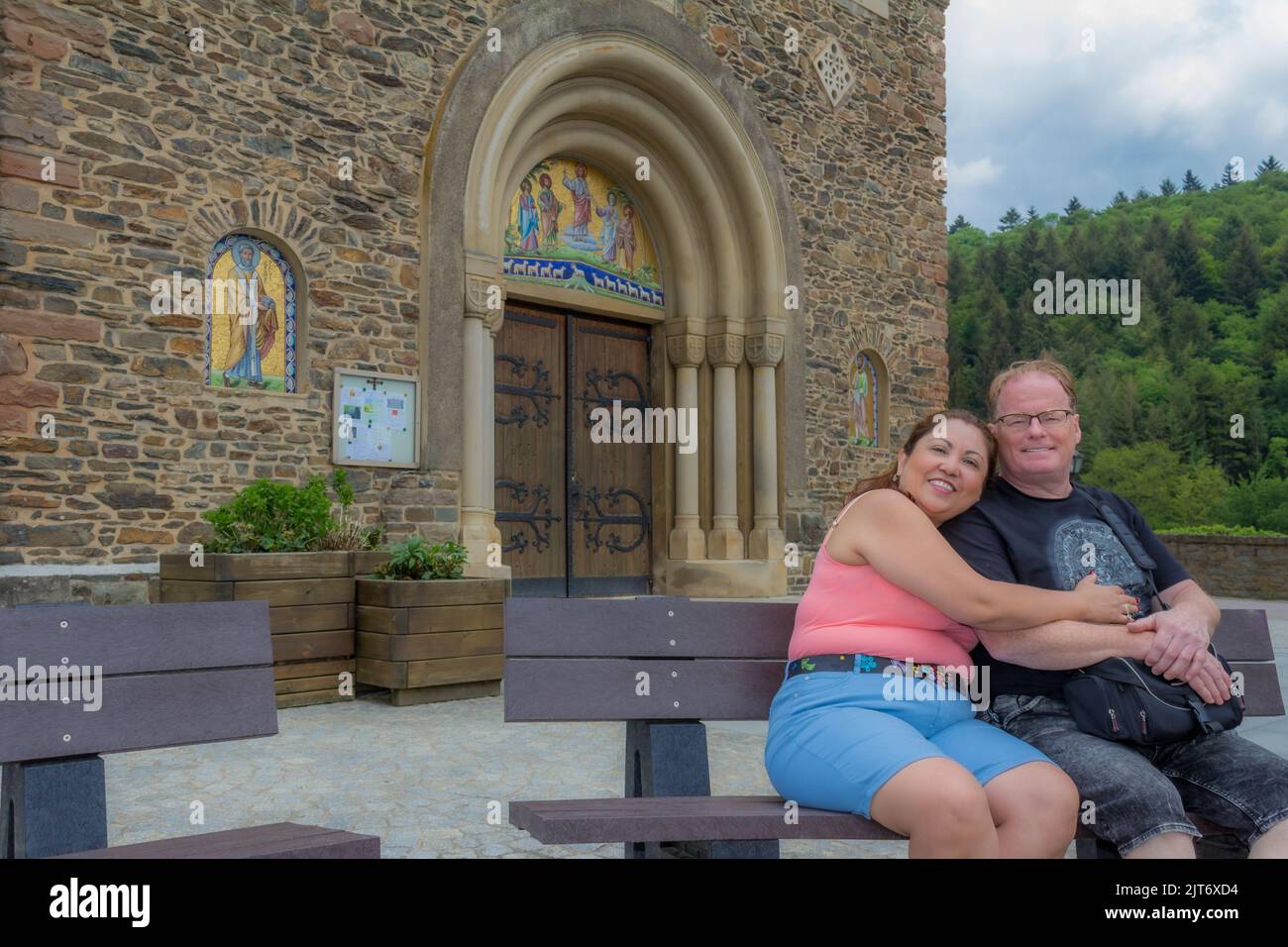 Lächelndes Touristenpaar mittleren Alters sitzt und umarmt auf Bank gegen Steinmauer und Eingang der Kirche Heiligen Cosmas und Damian, Bäume im Rücken Stockfoto