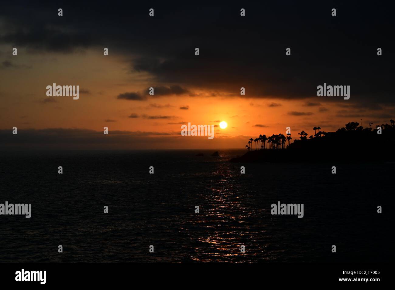 Ein strahlend orangefarbener Himmel, wenn die Sonne zwischen Wolkenschichten über Twin Points, Laguna Beach, Kalifornien, untergeht. Stockfoto