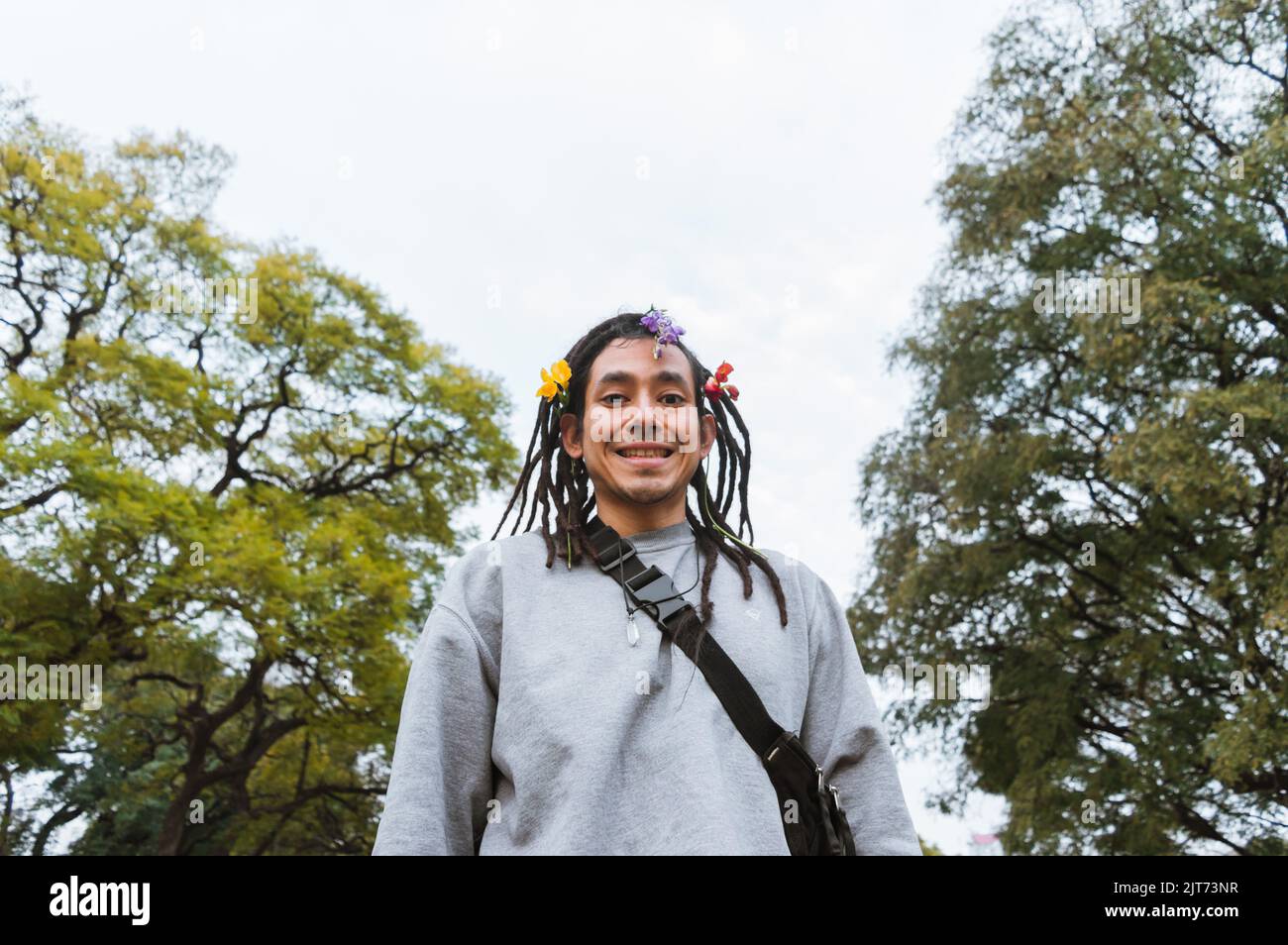 lateinischer venezolanischer junger Mann mit Dreadlocks und Blumen auf den Haaren steht schüchtern und lächelnd im Park und blickt auf die Kamera, mit Bäumen und dem Himmel Stockfoto