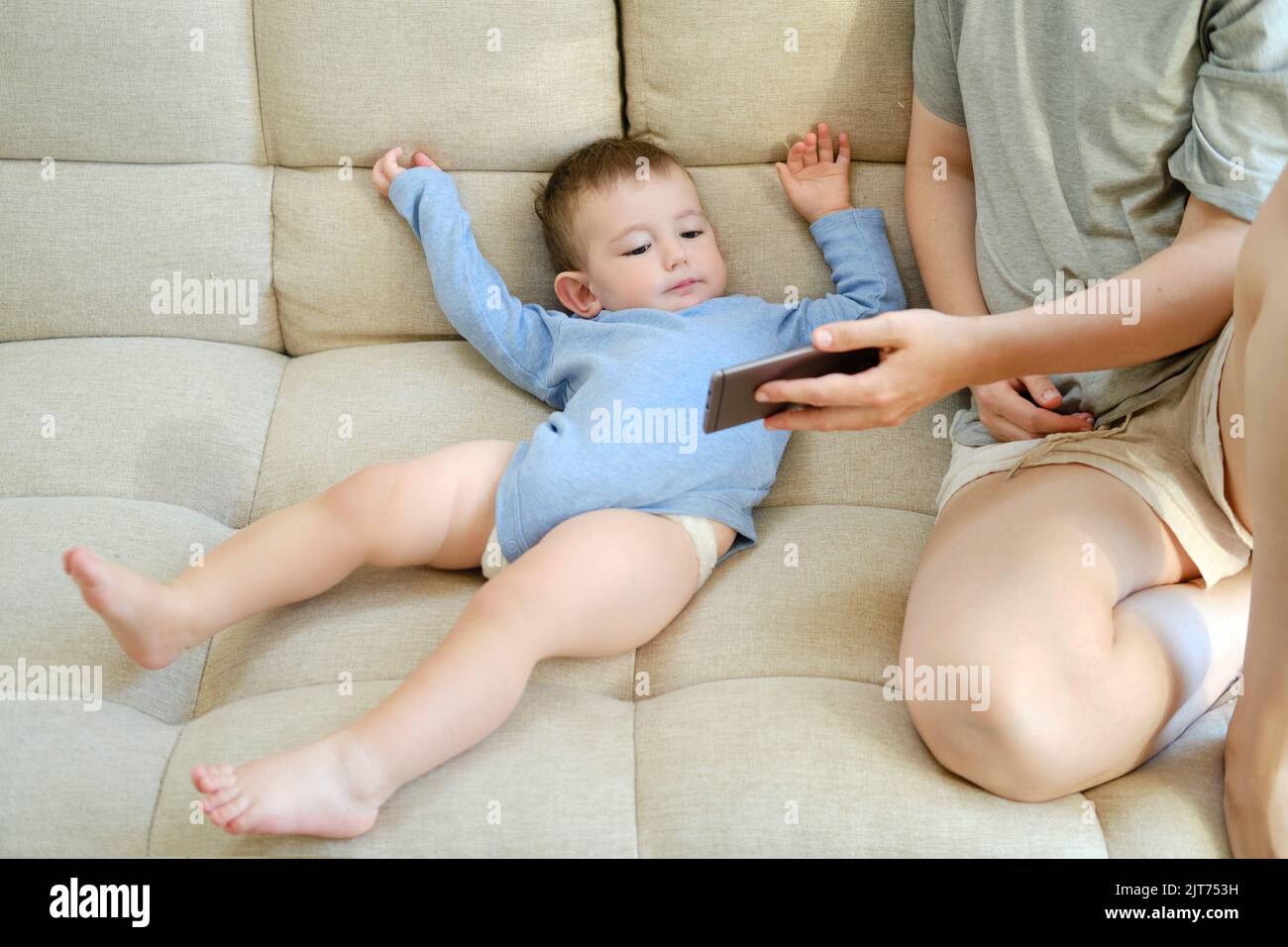 Kleinkind Baby sitzt mit seiner Mutter und schaut in das Telefon. Mutter und Kind mit einem Smartphone in den Händen auf dem Sofa Stockfoto