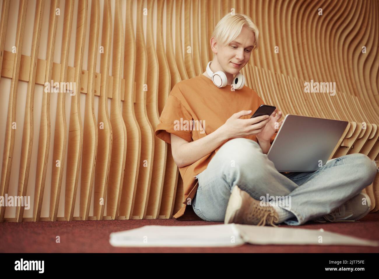 Glücklicher blonder Teenager in der Freizeit, der in einem Smartphone schreibt, während er in der Pause oder nach dem Unterricht auf dem Boden des College-Korridors sitzt Stockfoto