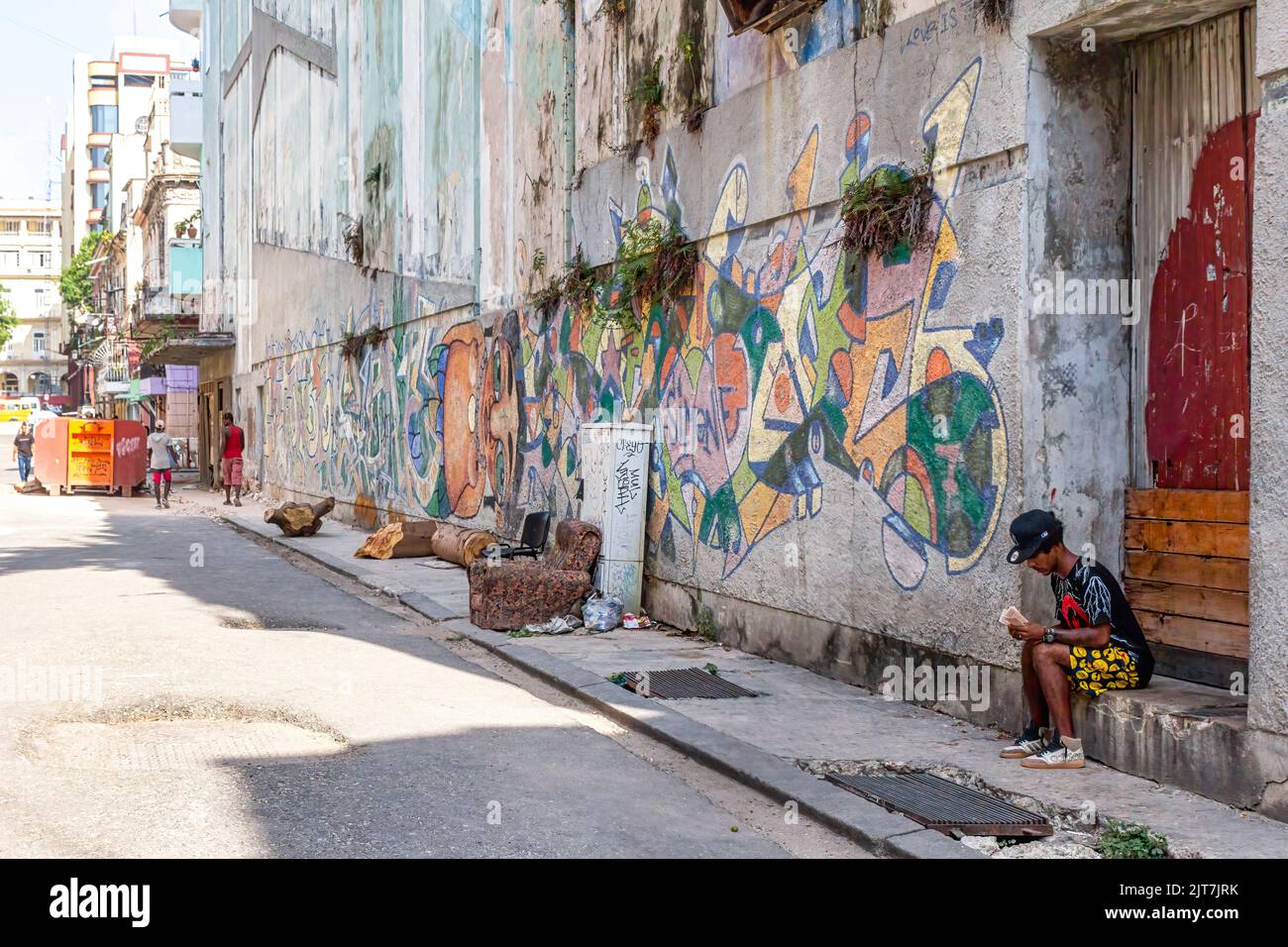 Ein Kubaner sitzt in einer Türschwelle auf einem schmutzigen und kaputten Bürgersteig. Die Gebäudewand hat städtische Graffiti. Stockfoto