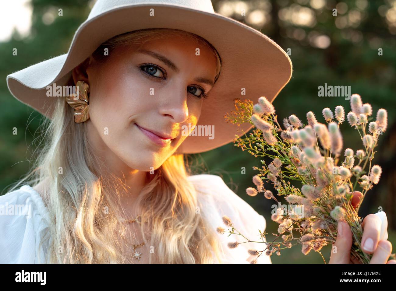 Porträt einer jungen, fröhlichen, atemberaubenden Frau, die goldene Ohrringe und einen beigen Schlapphut trägt und bei Sonnenuntergang im Waldpark steht. Stockfoto