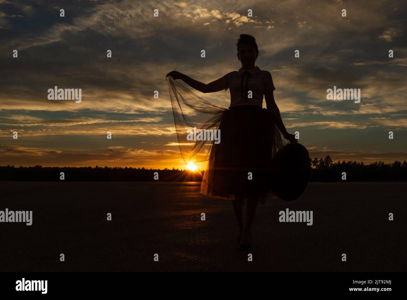 Low-Winkel-Ansicht der Märchenfrau halten Kante des Tutu Rock durch die Sonne gegen Wald Silhouette und Sonnenuntergang bewölkten Himmel mit Hut in der Hand hinterleuchtet Stockfoto