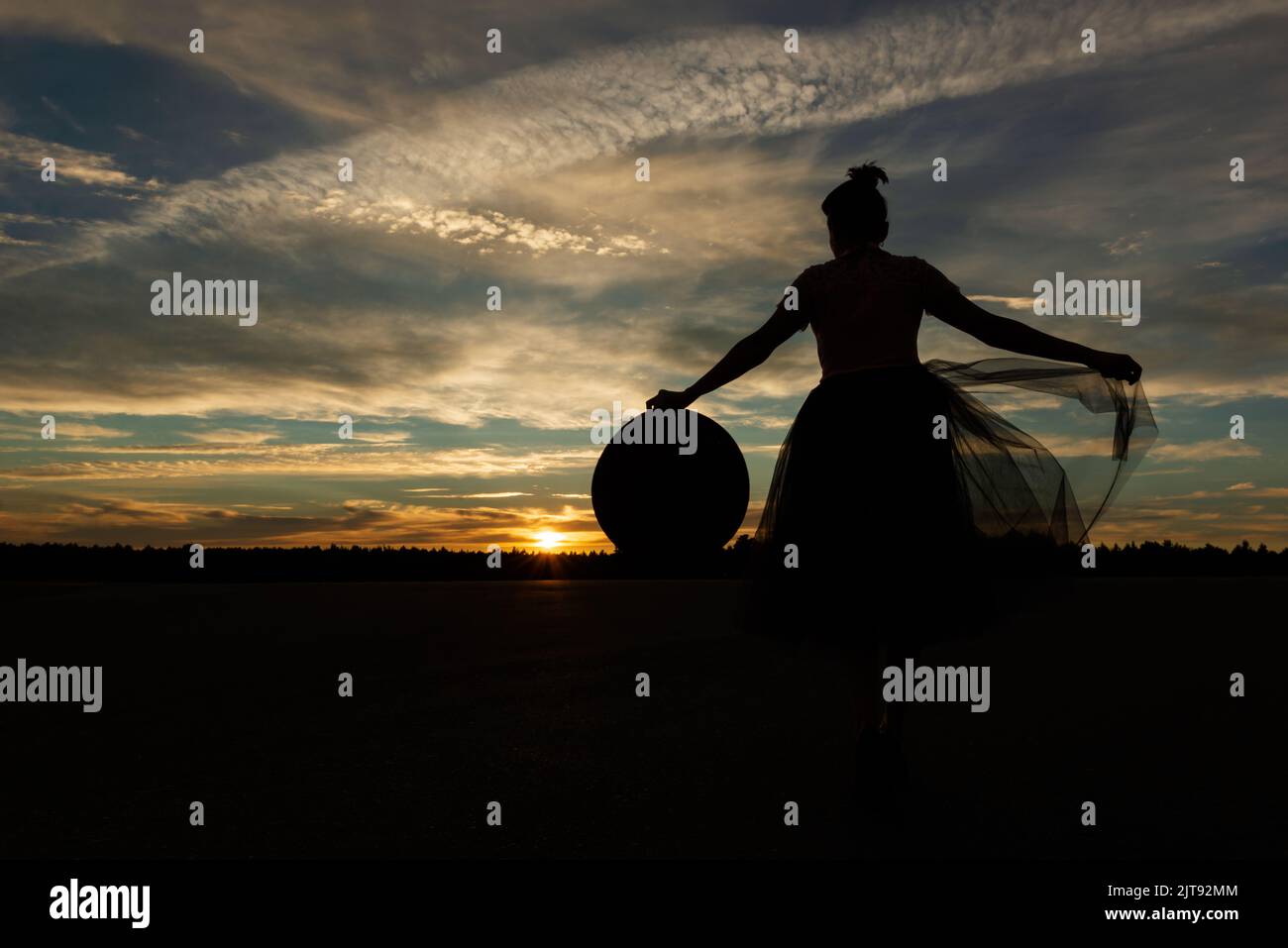 Silhouette einer märchenhaften Frau, die gegen den Sonnenuntergang steht, mit ausgestreckten Armen, die ihren Hut und den Rand des Tutu-Rockes gegen den Wald und den wolkigen Himmel hält Stockfoto