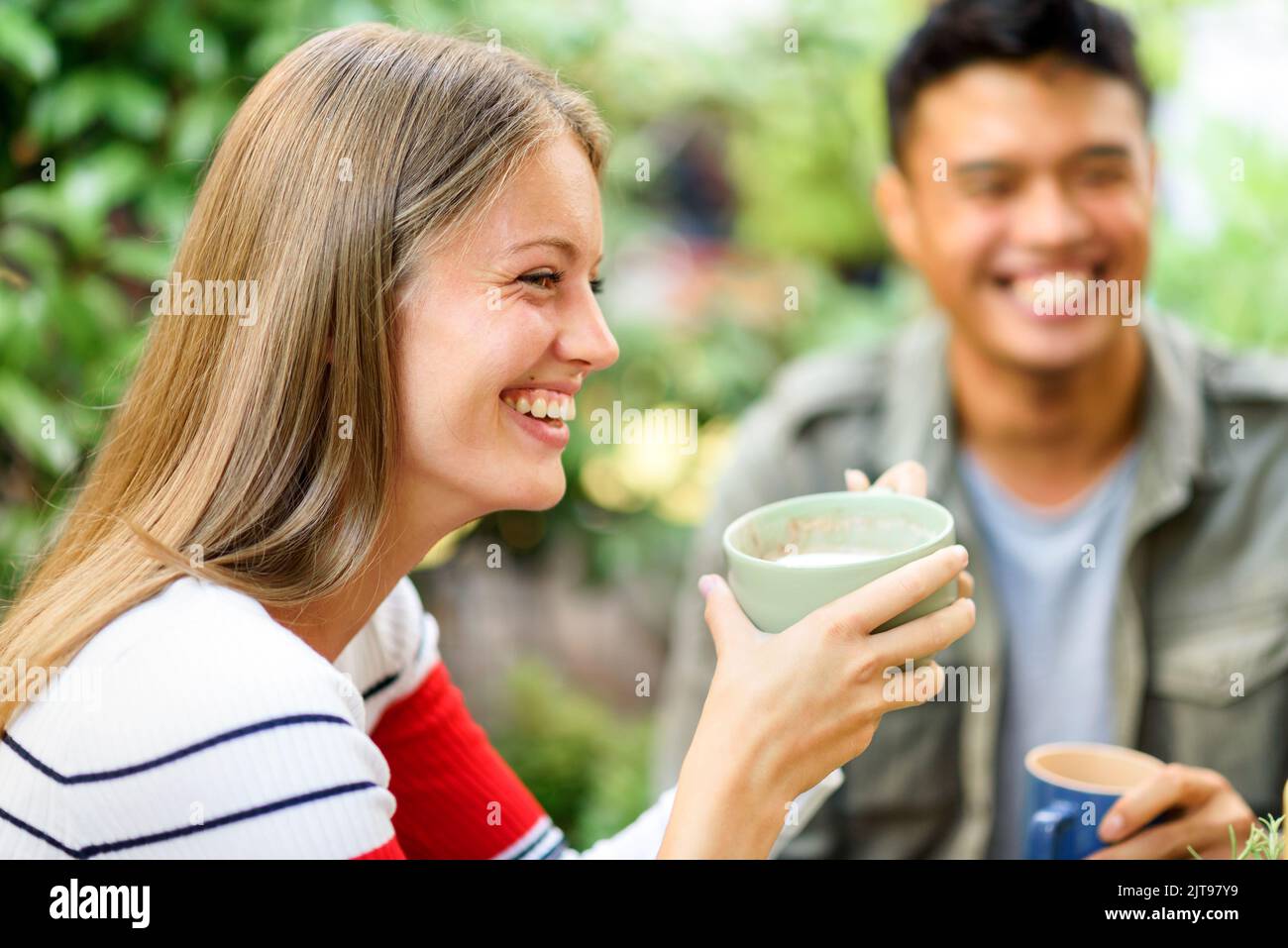 Positive Frau mit einer Tasse Cappuccino auf der Terrasse in der Nähe eines asiatischen Mannes auf verschwommenem Hintergrund beim Frühstück am Sommermorgen Stockfoto