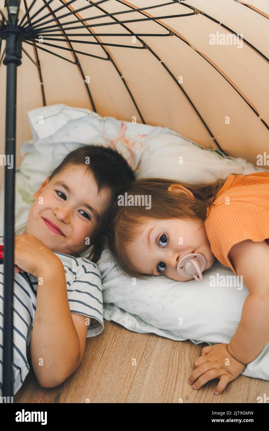 Kinder im Pyjama spielen zu Hause und liegen auf dem Boden unter einem Regenschirm, versteckt vor ihren Eltern. Ruhen auf Heizboden. Junge und Mädchen posieren für Stockfoto